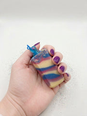 A hand holding a blackberry sippy soap bar with blue, pink and cream swirls and a charm topper